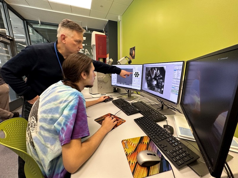 Two people working at a desk with three computer monitors displaying technical data.