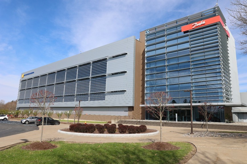A modern office building with glass windows and a metal facade, featuring a red sign and landscaped surroundings.