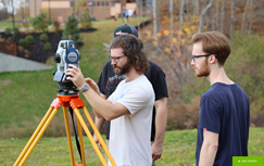 Three individuals surveying outdoors; one adjusting a surveying instrument on a tripod, with autumn foliage in the background.